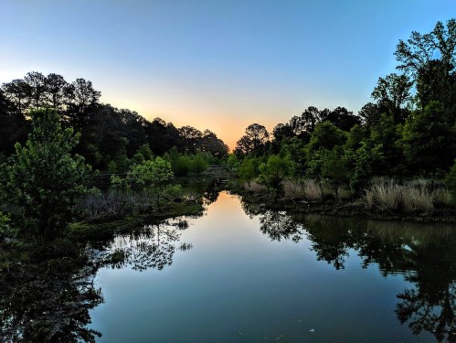 A serene landscape featuring a calm pond reflecting trees and a colorful sunset sky.