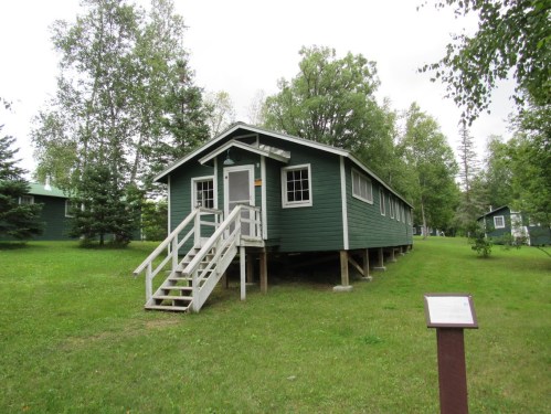 A green wooden cabin on stilts surrounded by trees and grass, with a set of stairs leading to the entrance.