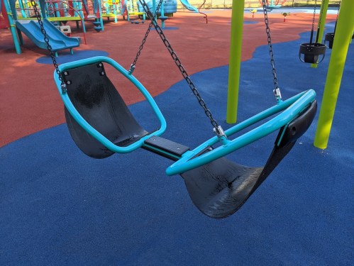 A double swing set with teal frames and black seats, hanging from chains in a colorful playground.