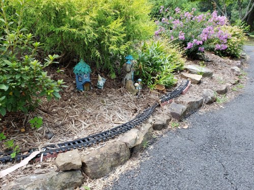 A garden scene featuring a miniature train track, a small house, and decorative figures among lush greenery and flowers.