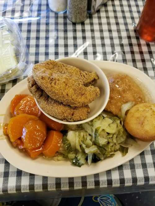 A plate of fried catfish, collard greens, peach slices, cornbread, and a side of gravy on a checkered tablecloth.