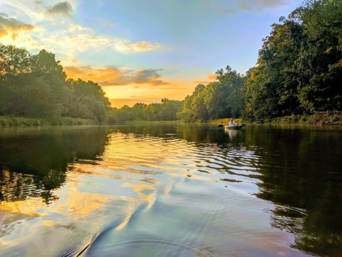 A serene river scene at sunset, with vibrant colors reflecting on the water and a lone person in a small boat.
