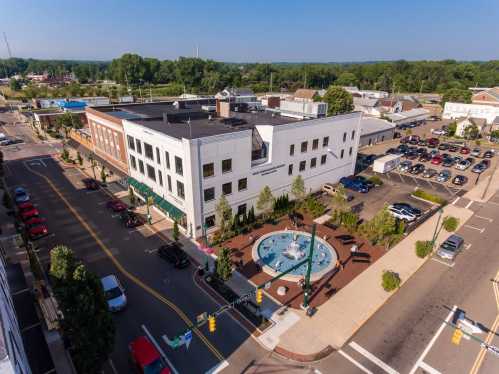 Aerial view of a town square featuring a fountain, surrounding buildings, and parked cars on a sunny day.