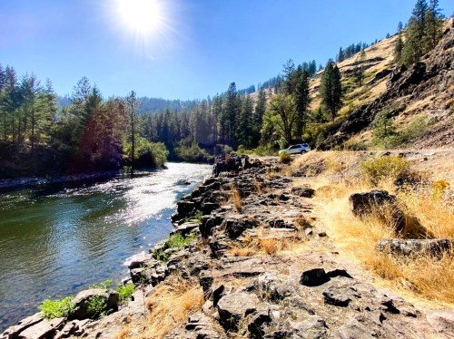 A sunlit riverbank with rocky shores, surrounded by trees and hills under a clear blue sky.