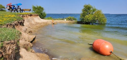 A calm lake scene with a buoy, grassy shore, and people sitting under umbrellas in the background on a sunny day.