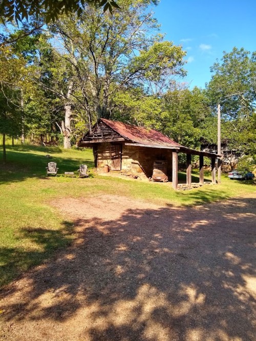 A rustic cabin with a weathered roof, surrounded by trees and a grassy area, with two chairs in front.