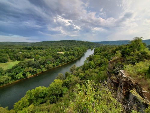 A scenic view of a winding river surrounded by lush green trees and rolling hills under a cloudy sky.