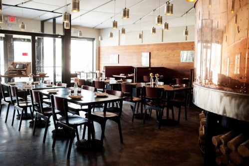 A modern restaurant interior with wooden accents, tables set for dining, and pendant lights hanging from the ceiling.
