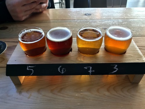A wooden flight board with four small glasses of different colored beers, labeled 5, 6, 7, and 3.