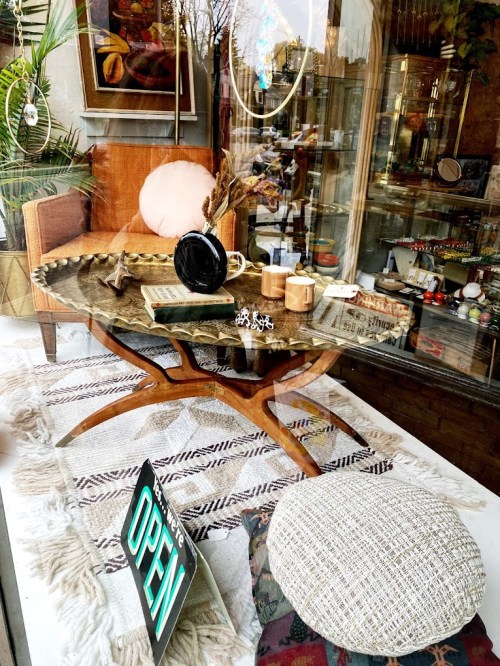 A cozy shop window display featuring a stylish table, armchair, decorative items, and an "OPEN" sign.