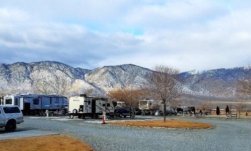 A scenic view of RVs parked near a gravel road, with snow-capped mountains and bare trees in the background.