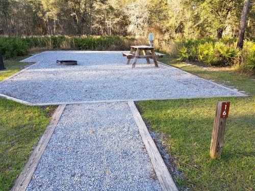 Gravel campsite with a picnic table, fire pit, and numbered post, surrounded by greenery and trees.
