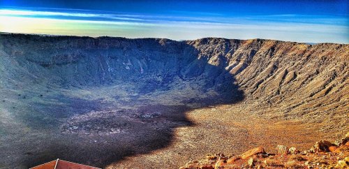 A vast, rocky crater with steep walls and a clear blue sky above, showcasing the impact site of a meteor.