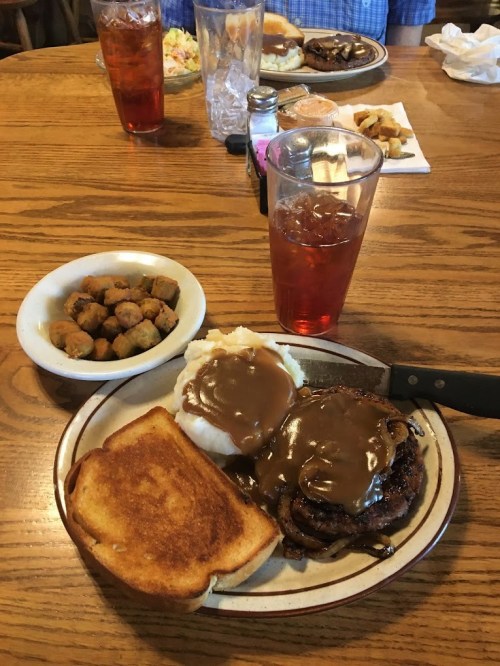 A plate with a meat patty topped with gravy, mashed potatoes, toast, and a side of okra, with iced tea in the background.