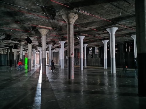 A spacious industrial interior with tall columns, polished concrete floors, and dramatic lighting creating long shadows.