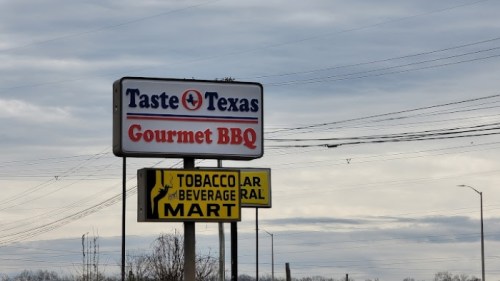 Sign for "Taste of Texas Gourmet BBQ" with a nearby "Tobacco & Beverage Mart" sign under a cloudy sky.