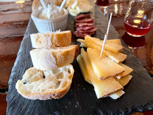 A slate platter with sliced bread, cheese, cured meats, and a small bowl of spread, accompanied by drinks.