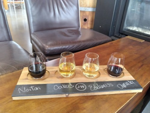 A wooden flight board with four glasses of wine: two red and two white, labeled with names on a chalkboard surface.
