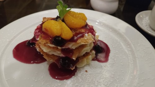 A layered dessert with crispy pastry, cream, berries, and citrus, garnished with mint on a white plate.