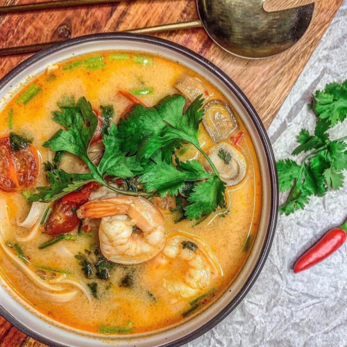 A bowl of flavorful shrimp soup garnished with fresh cilantro, surrounded by herbs and a chili pepper on a wooden surface.