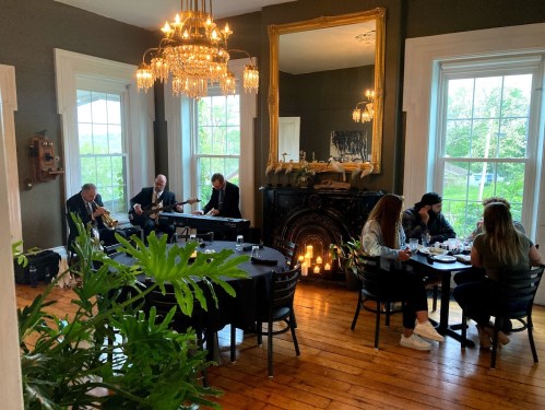 A cozy dining room with live musicians playing, guests enjoying meals, and a warm ambiance from candles and greenery.