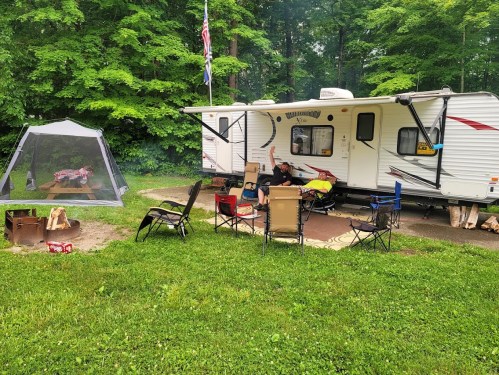 A campsite with a trailer, chairs, a fire pit, and a screened tent surrounded by lush green trees.