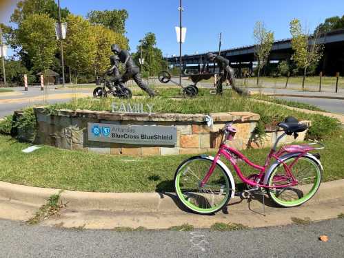 A pink bicycle rests beside a stone monument featuring sculptures of cyclists and the word "Family."