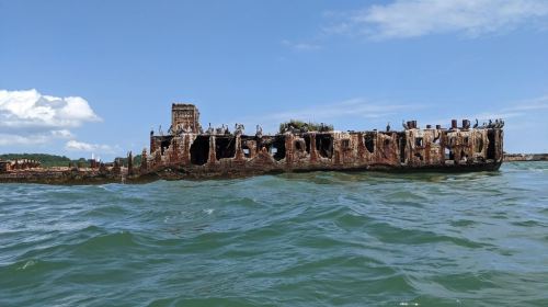 A rusted shipwreck partially submerged in water, with visible holes and greenery on top, under a blue sky.