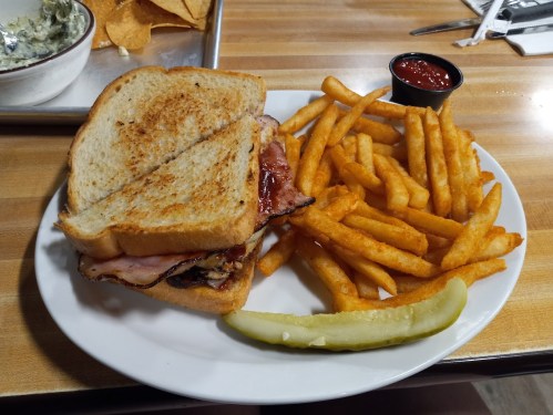 A plate with a toasted sandwich filled with ham, served with golden fries and a pickle, alongside a small cup of ketchup.