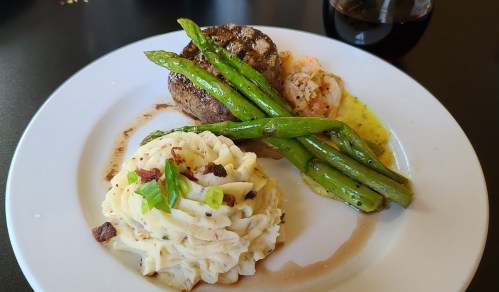 A plate featuring grilled steak, asparagus, shrimp, and creamy pasta with green onions, accompanied by a glass of red wine.