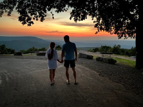 A couple holds hands while walking on a path at sunset, surrounded by trees and mountains in the background.