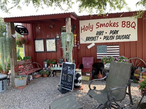 A rustic BBQ stand with a sign reading "Holy Smokes BBQ," featuring menu items and a cozy outdoor seating area.