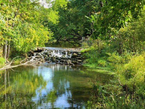 A serene landscape featuring a small waterfall over rocks, surrounded by lush green trees and calm water reflections.