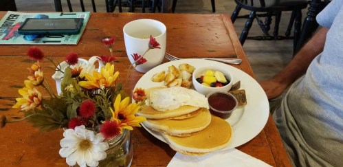 A plate of pancakes with a poached egg, fruit, and potatoes, alongside a cup of tea and a flower vase on a wooden table.