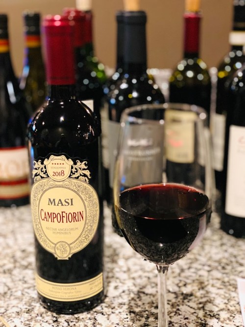 A bottle of Masi Campofiorin 2018 red wine next to a filled wine glass, with various wine bottles in the background.