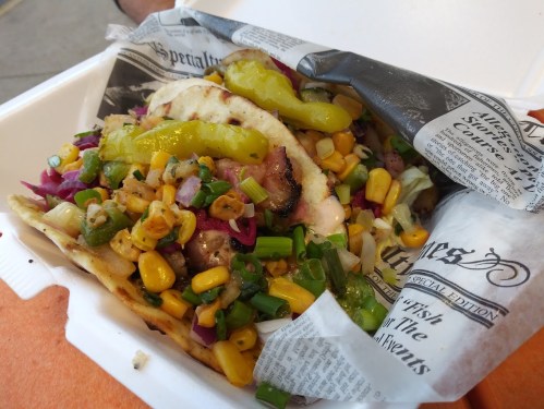 A taco filled with grilled meat, corn, onions, and peppers, served in a takeout container lined with newspaper.