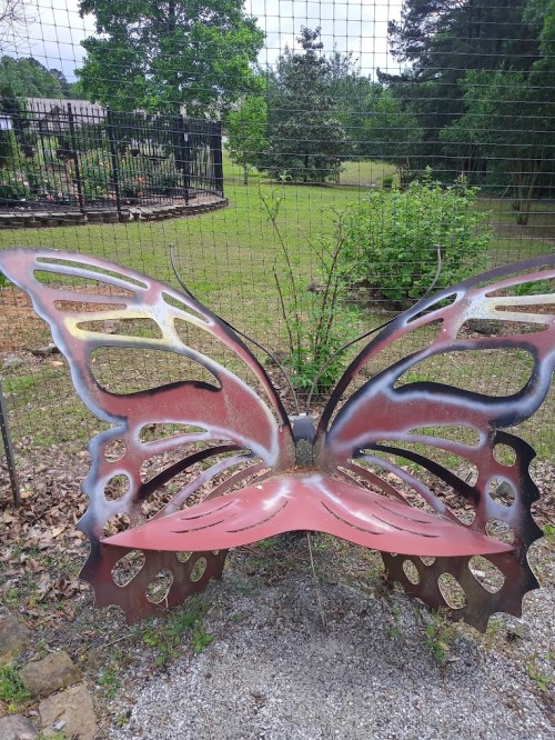 A large, decorative butterfly-shaped bench in a garden, surrounded by greenery and a fence in the background.