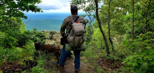 A person in camouflage stands on a trail with a dog, overlooking a scenic view of mountains and greenery.