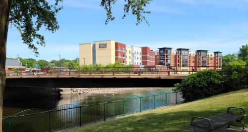 A bridge over a river with a modern hotel in the background, surrounded by greenery and blue skies.