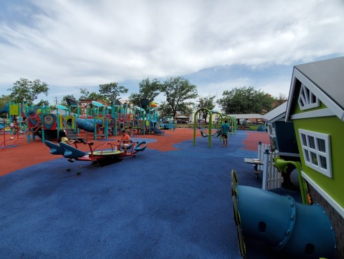 A colorful playground with slides, swings, and play structures, surrounded by trees and a blue sky.