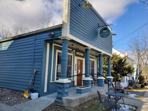 A blue wooden building with a sign, featuring a porch and outdoor seating, under a partly cloudy sky.