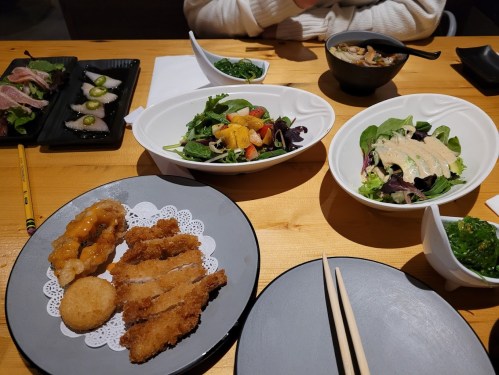 A table set with various dishes: fried chicken, salad, sashimi, and a bowl of soup, with chopsticks and napkins.