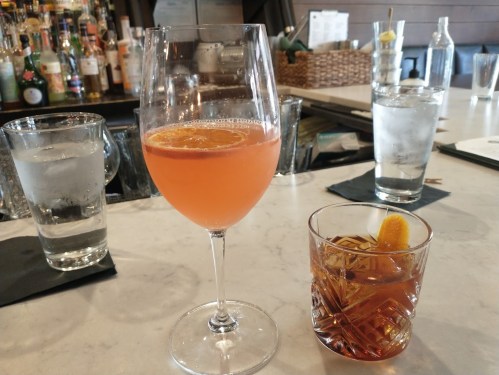 A glass of pink cocktail with an orange slice and a short glass of amber drink on a marble bar counter.