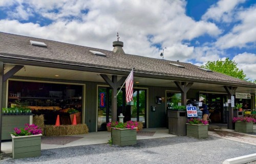 A rustic storefront with an American flag, flower planters, and an "Open" sign under a partly cloudy sky.