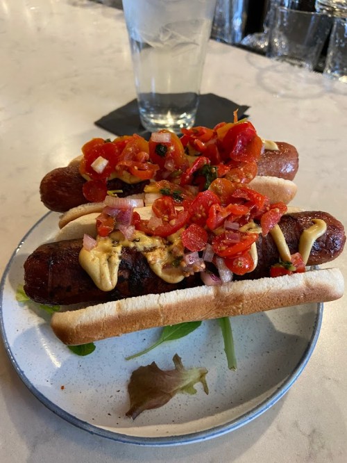 A plate with three hot dogs topped with diced tomatoes, onions, and mustard, alongside a glass of water.