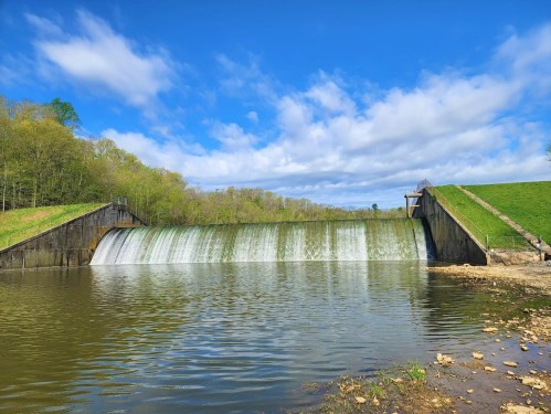 A serene landscape featuring a dam with water cascading over it, surrounded by greenery and a blue sky.