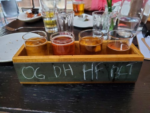 A wooden flight tray with four small glasses of different colored beverages, labeled "OG DH HF BLT."