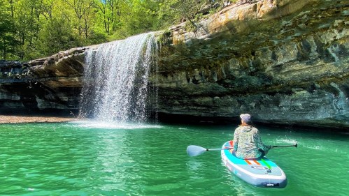 A person paddles on a kayak in clear water, facing a waterfall surrounded by lush greenery.