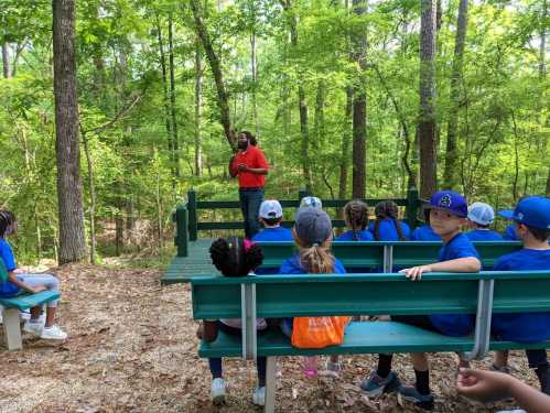 A group of children in blue shirts sits on benches in a forest, listening to a speaker in a red shirt.