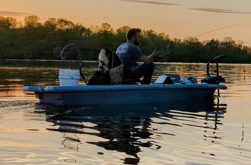 A person fishing from a small boat on a calm lake during sunset, surrounded by trees and reflecting water.